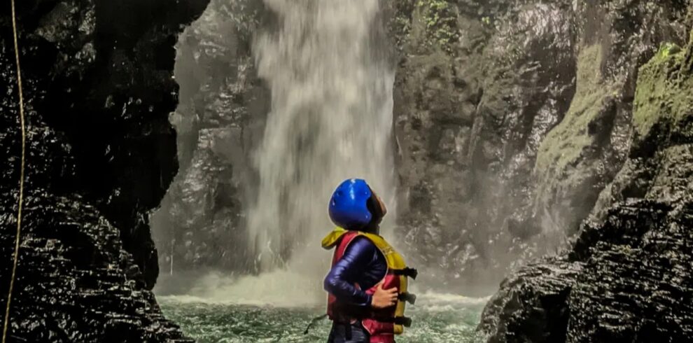 Solo adventurer standing in the water beneath Curug Naga waterfall in Bogor, West Java, wearing safety gear while surrounded by towering canyon walls, cascading water, and lush tropical rainforest.