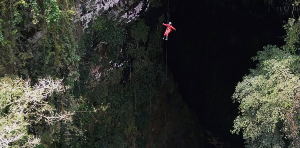 A caver in a red jumpsuit rappels into the vast vertical entrance of Goa Ngeleng in Yogyakarta, Indonesia. Surrounded by lush green vegetation, limestone cliffs, and a dramatic underground sinkhole, the adventurer descends into one of Jogja's most impressive cave systems.