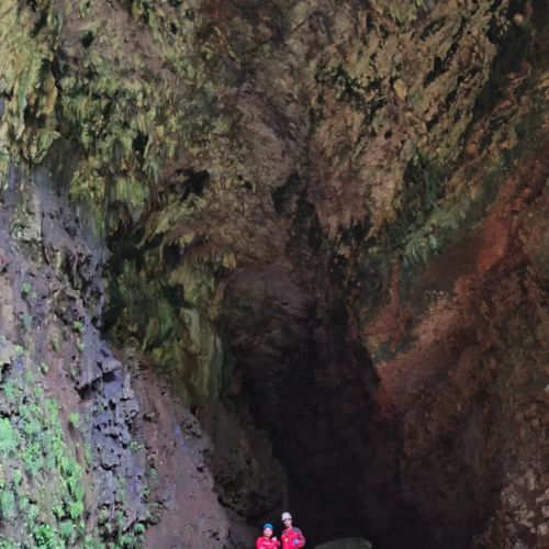 Two cavers wearing red safety suits and helmets stand inside the enormous chamber of Goa Ngeleng in Gunungkidul, Yogyakarta, Indonesia. Surrounded by towering limestone walls, ancient cave formations, and a vast underground cavern, the explorers showcase the immense scale of this spectacular natural wonder.