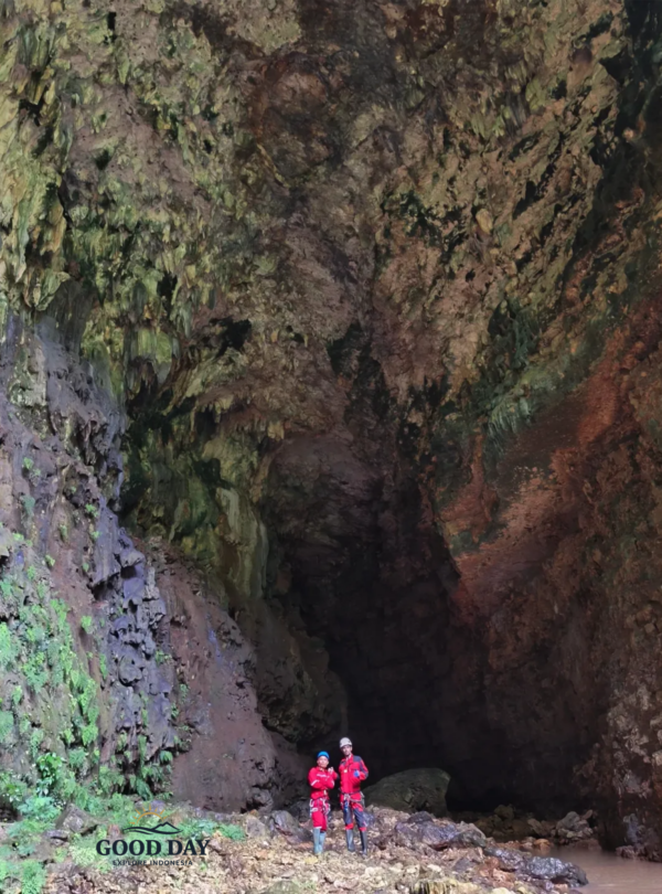 Two cavers wearing red safety suits and helmets stand inside the enormous chamber of Goa Ngeleng in Gunungkidul, Yogyakarta, Indonesia. Surrounded by towering limestone walls, ancient cave formations, and a vast underground cavern, the explorers showcase the immense scale of this spectacular natural wonder.