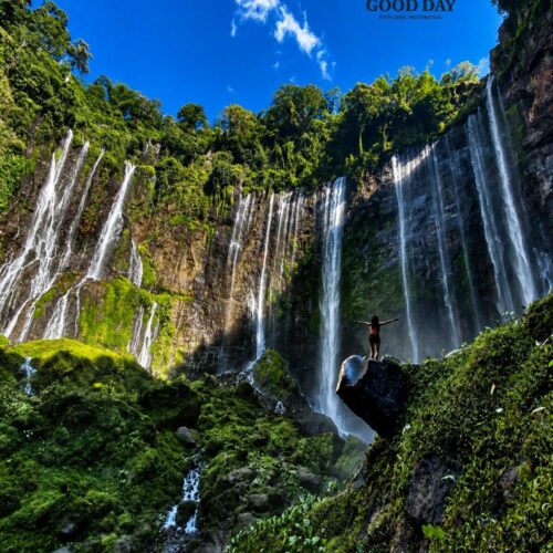 A traveler standing on a rock with arms outstretched in front of Tumpak Sewu Waterfall, surrounded by lush green cliffs, cascading waterfalls, tropical vegetation, and a vibrant blue sky in East Java, Indonesia
