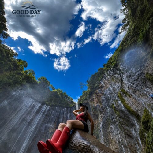 A traveler wearing red boots sits on a large rock beneath the towering Tumpak Sewu Waterfall in East Java, Indonesia, surrounded by lush cliffs, cascading waterfalls, tropical rainforest, and a dramatic blue sky with clouds overhead.