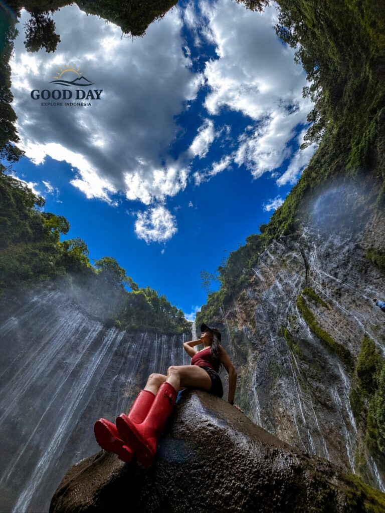 A traveler wearing red boots sits on a large rock beneath the towering Tumpak Sewu Waterfall in East Java, Indonesia, surrounded by lush cliffs, cascading waterfalls, tropical rainforest, and a dramatic blue sky with clouds overhead.