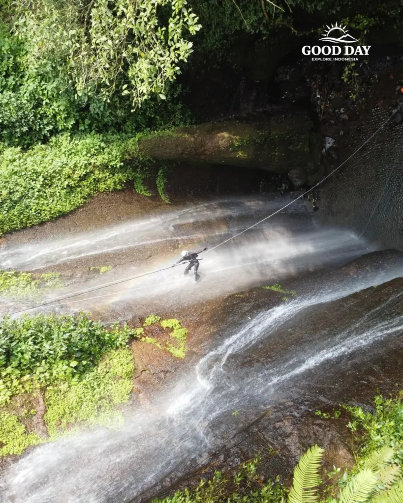 A canyoning participant rappelling down Curug Nangka Waterfall in Bogor, Indonesia, surrounded by lush rainforest during a guided adventure by Good Day.